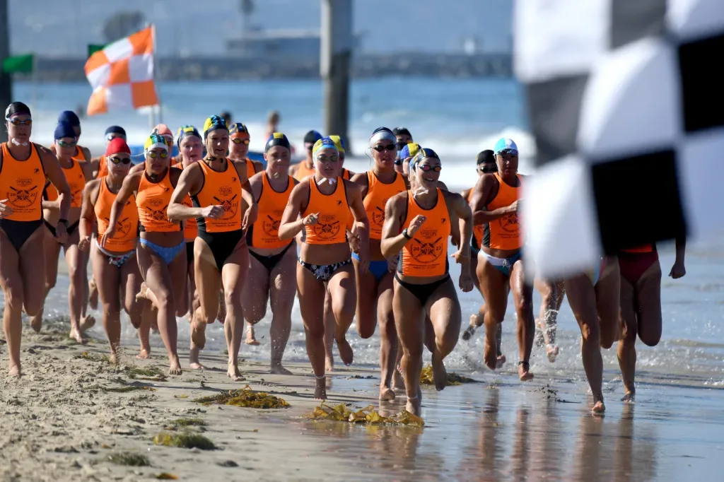 Ocean swimmers in orange tank tops and swim caps sprinting out of the surf at the start of a beach race in the South Bay, Los Angeles