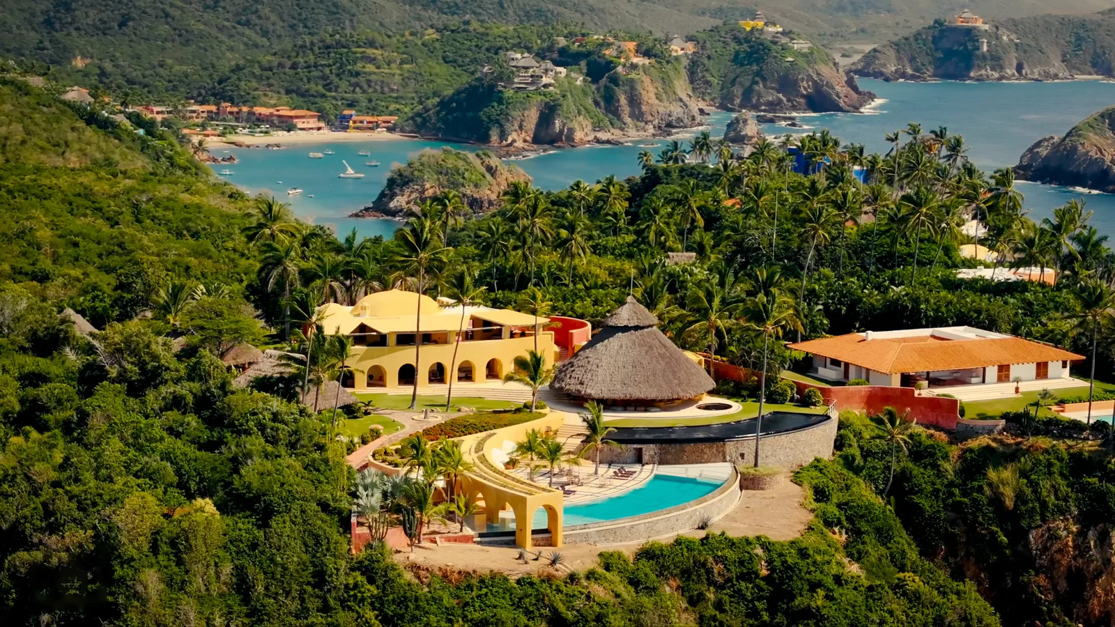 Aerial view of a yellow Mexican villa with a thatched palapa roof and a curved infinity pool set into lush palm jungle above a turquoise Pacific cove on Mexico's Pacific coast