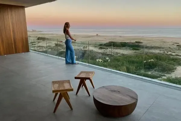 Minimalist concrete-and-glass terrace with cane stools and a round wood table at sunset, overlooking grassy dunes and a wide Atlantic beach on the Uruguayan coast beyond Punta del Este
