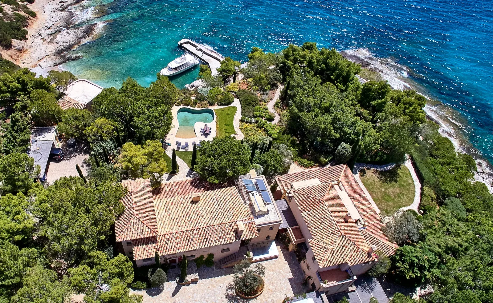 Aerial view of a Mediterranean stone villa with terracotta-tiled roofs, a garden pool, cypress trees and a private dock on turquoise water on the Peloponnese coast, Greece