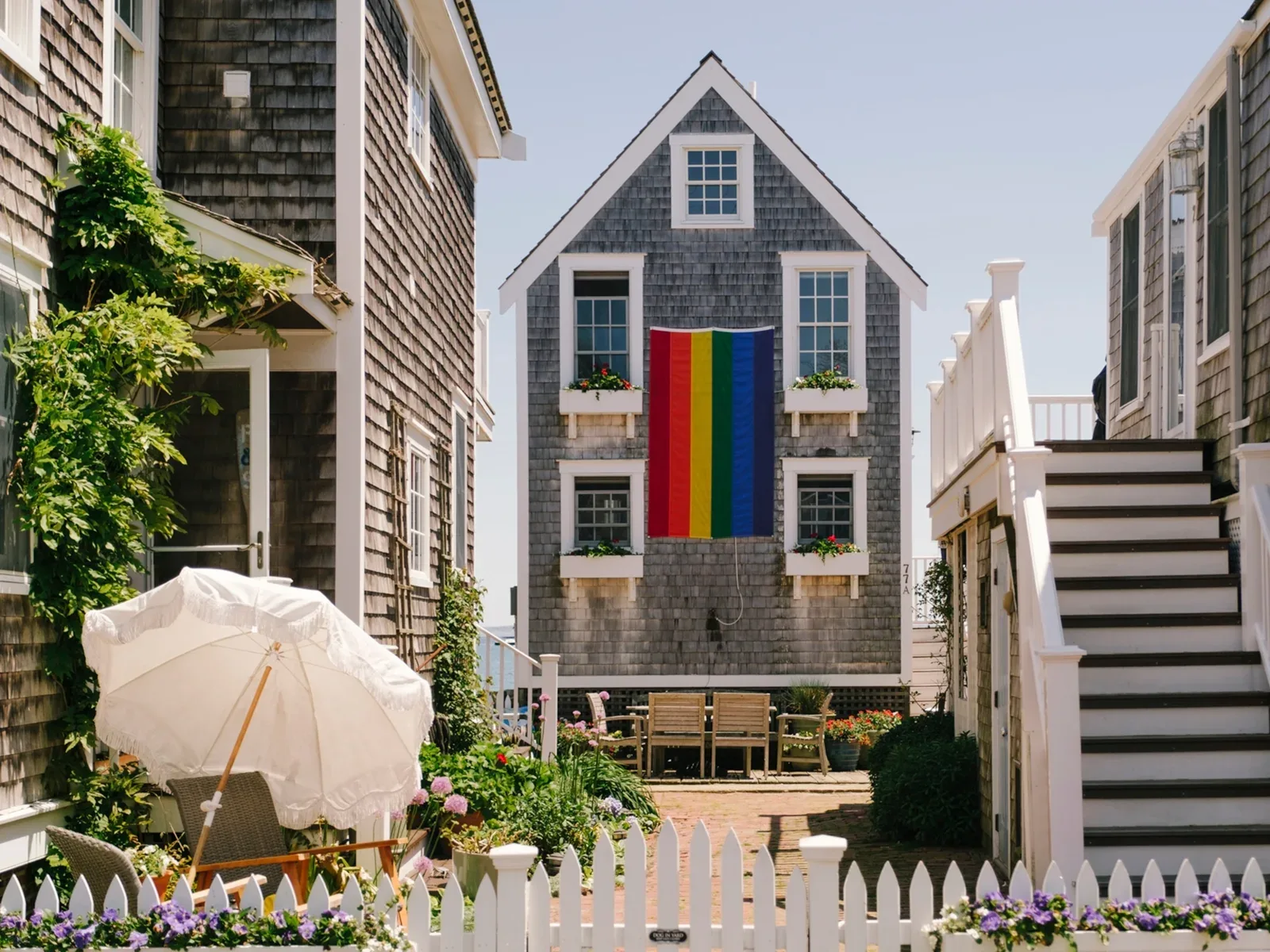 Grey-shingled Cape Cod cottage in Provincetown flying a large vertical rainbow pride flag, behind a white picket fence and flower garden