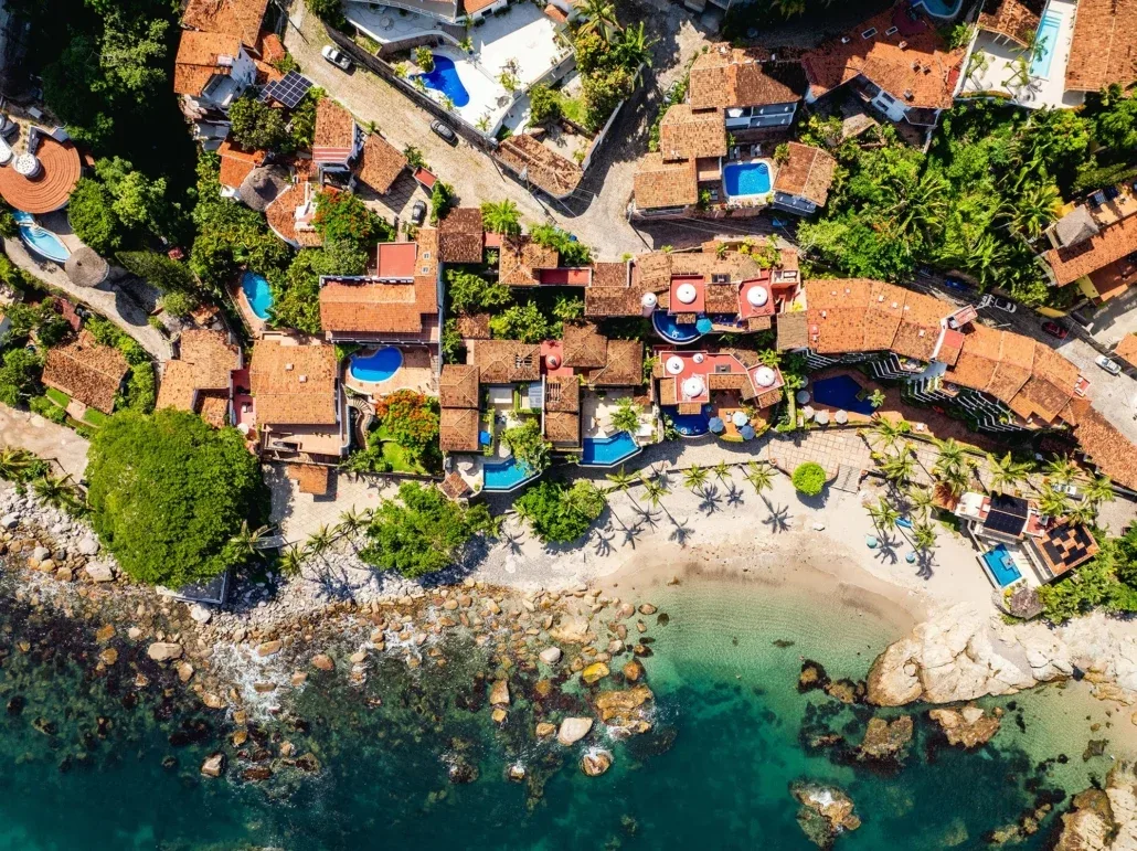 Aerial view of terracotta-roofed hillside villas with private pools above a small sandy cove and turquoise sea in Puerto Vallarta, Mexico