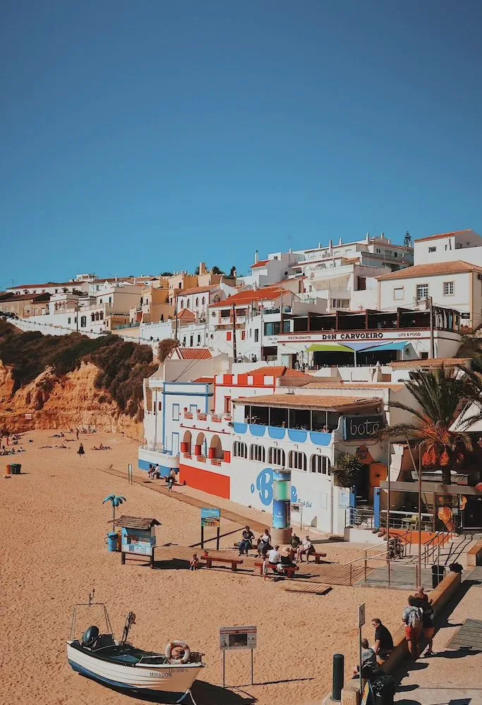 Golden-sand beach cove at Carvoeiro in the Algarve, Portugal, backed by ochre cliffs and a hillside of whitewashed, terracotta-roofed buildings, with a small fishing boat on the sand under a clear blue sky