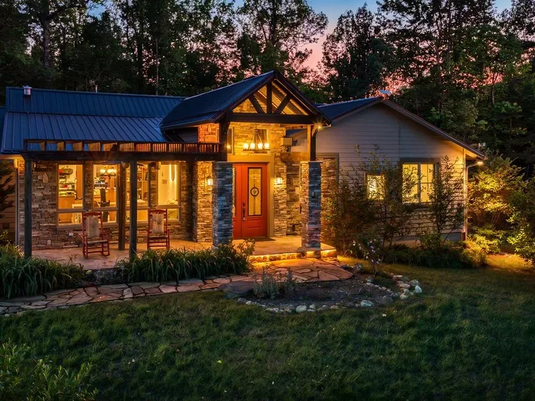Stone-and-timber craftsman mountain home at dusk with a lit porch and metal roof, set in the forested hills near Asheville, North Carolina