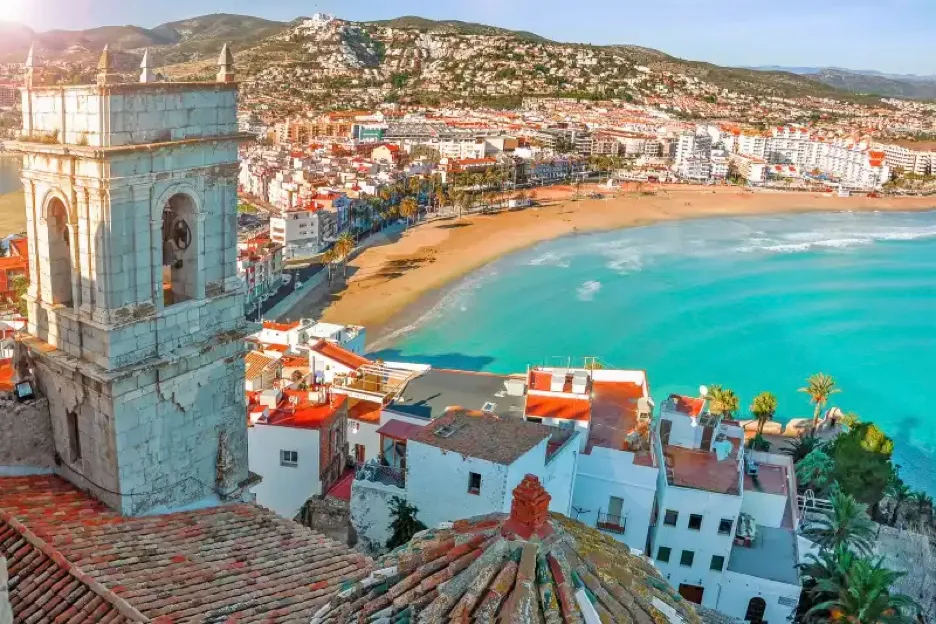 Sunlit Valencian coastal town with a historic stone bell tower overlooking a curved golden-sand bay of turquoise water, terracotta-roofed whitewashed houses on the hillside and green mountains behind