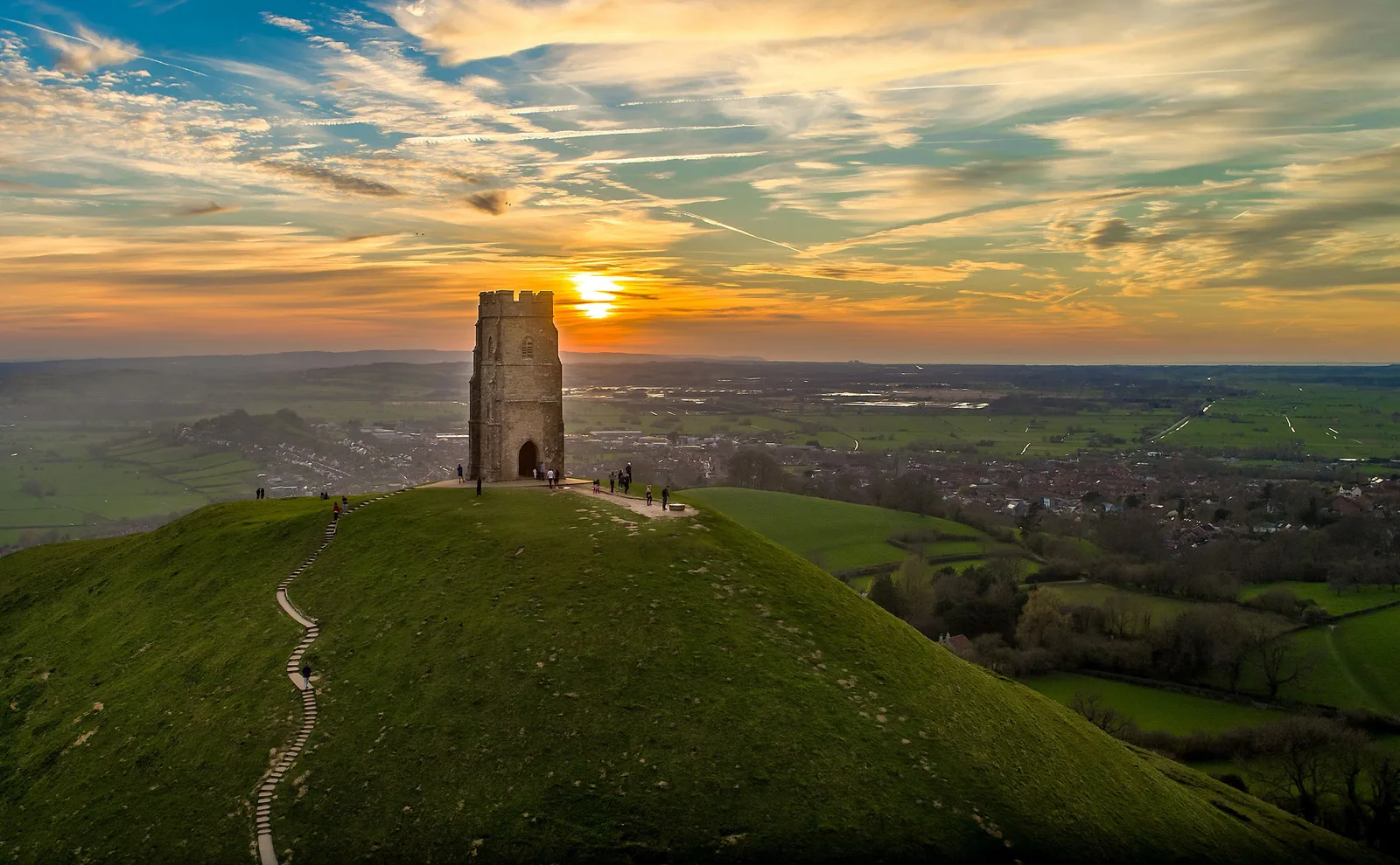Aerial view of St Michael's Tower atop the grassy hill of Glastonbury Tor at golden sunset over the Somerset Levels, England