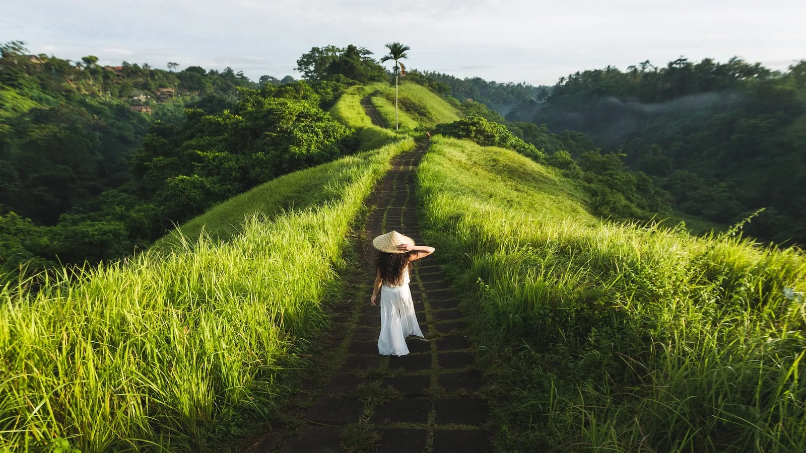 A woman in a white dress and conical straw hat walking the Campuhan Ridge Walk path between lush green ridges at sunrise near Ubud, Bali