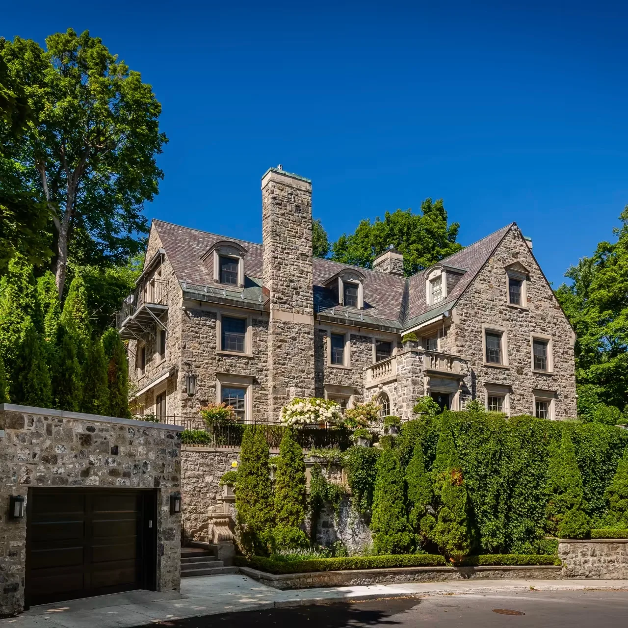 Grand grey-stone heritage mansion with a slate roof, gabled dormers and mature hedges and trees, in the Westmount district of Montreal, Canada