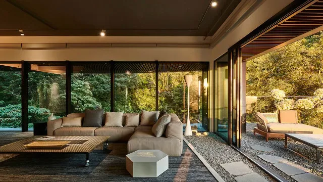 Modern living room with sliding glass walls opening onto a lush forested hillside at dusk, in the green hills around Taipei, Taiwan